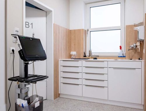 Blick in ein Laborzimmer in einer Arztpraxis. Das Labor oder Untersuchungszimmer hat weiße Unterschränke, eine Wandverkleidung aus hellem Holzund viele Schubladen. Alles ist sehr hell und freundlich. Links im Raum steht ein Ultraschallgerät.