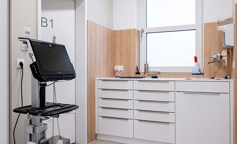 Blick in ein Laborzimmer in einer Arztpraxis. Das Labor oder Untersuchungszimmer hat weiße Unterschränke, eine Wandverkleidung aus hellem Holzund viele Schubladen. Alles ist sehr hell und freundlich. Links im Raum steht ein Ultraschallgerät.